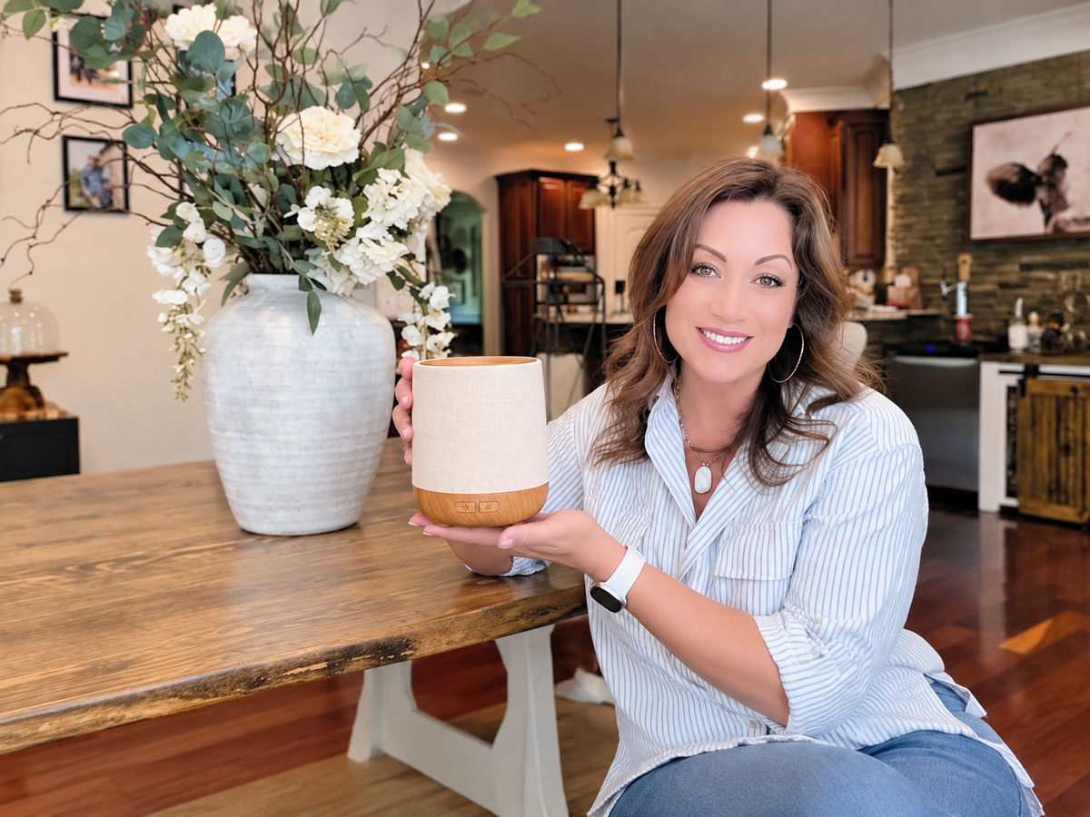 Smiling woman at a wooden dining table holding a ceramic and wood diffuser, one of our diffuser gift ideas for mom