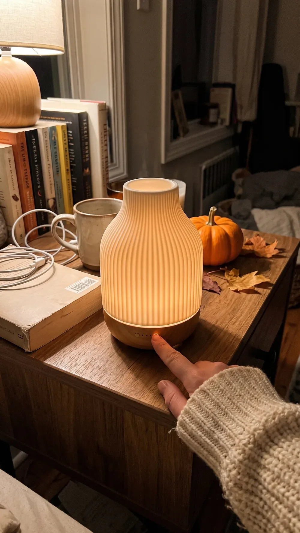A hand on the glowing ceramic diffuser next to a book and a pumpkin